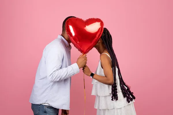Молодая романтическая чёрная пара целуется, прячась за воздушным шаром с красным сердцем — стоковое фото