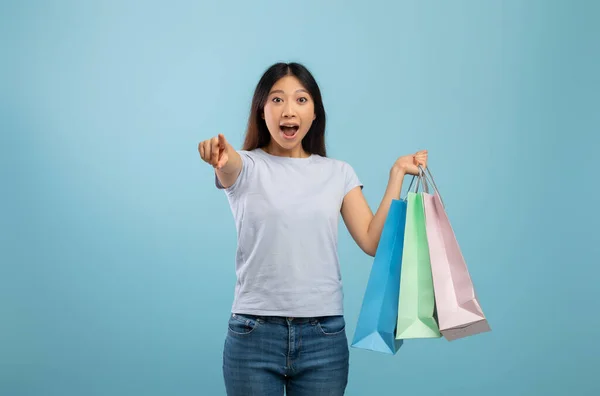 Ei, você. Surpreendido asiático senhora segurando coloridos sacos de compras e apontando para a câmera, posando sobre fundo azul — Fotografia de Stock