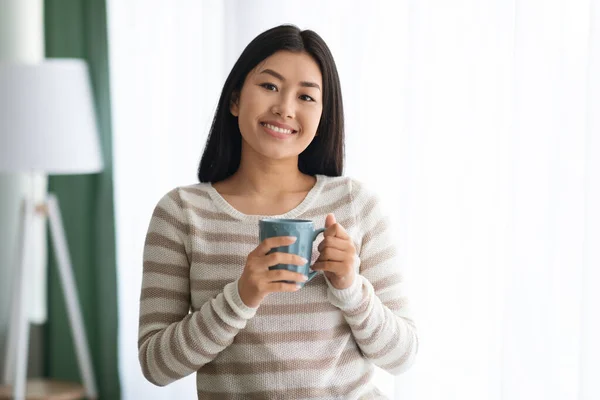 Mooie jonge aziatische vrouw drinken koffie terwijl staande in de buurt van venster thuis — Stockfoto