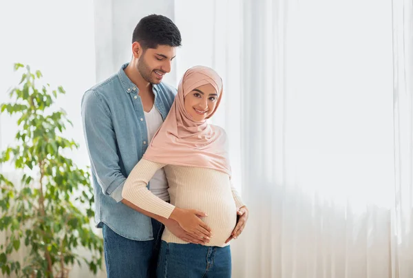 Los futuros padres. Embarazada musulmana mujer en hijab y su marido abrazándose junto a la ventana, hombre abrazando esposas vientre — Foto de Stock