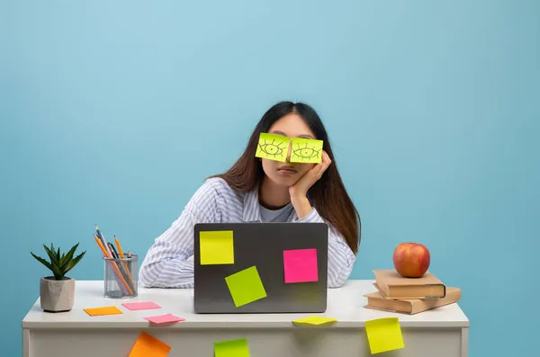 En utmattad elev. Porträtt av överarbetad dam sitter vid skrivbordet med klistermärken på ögonen och använda laptop, blå bakgrund — Stockfoto