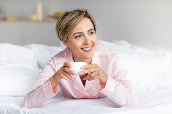 Ευτυχισμένη γυναίκα κοιτάζει αλλού και σκέφτεται, πίνοντας καφέ στο κρεβάτι — Φωτογραφία Αρχείου