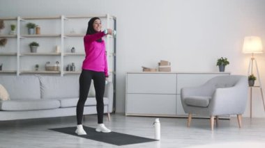 Aktif ortadoğulu genç bir kadın evde dambıllarla egzersiz yapıyor, sabahları antrenmanların keyfini çıkarıyor.