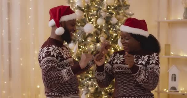 Rodinný vánoční večírek. Mladý africký Američan muž a žena nosí Santa klobouky tanec v blízkosti vánočního stromu doma — Stock video