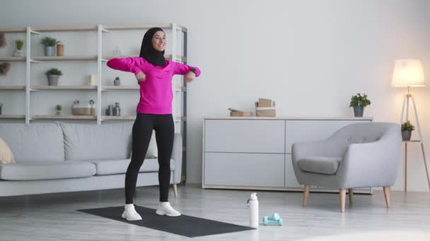 Salle de gym domestique. Jeune femme musulmane positive portant des vêtements de sport et du hijab faisant de l'exercice à la maison, réchauffant ses mains — Video