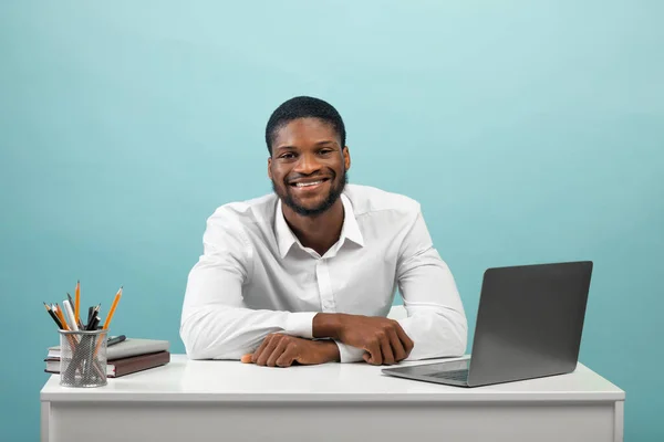 Retrato de homem freelancer afro-americano sentado na mesa com laptop, sorrindo para a câmera sobre fundo azul — Fotografia de Stock