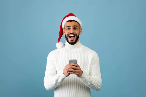 Chico árabe sorprendido en el teléfono inteligente de la celebración del sombrero de santa, consiguió oferta en línea móvil o mensaje de saludo de Navidad — Foto de Stock