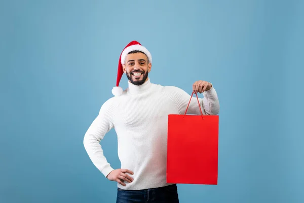Hombre árabe feliz con sombrero de Santa y bolsa de compras con regalo de Navidad sobre fondo azul, toma de estudio — Foto de Stock