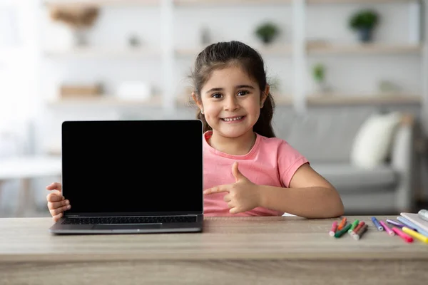 Sorrindo menina apontando para o laptop com tela preta em branco em casa — Fotografia de Stock