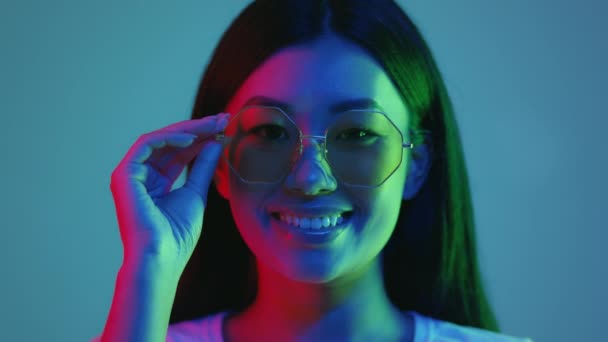 Neon ilumina as pessoas. Positivo asiático senhora modelo em óculos posando em luzes de néon brilhantes, sorrindo sobre fundo azul — Vídeo de Stock