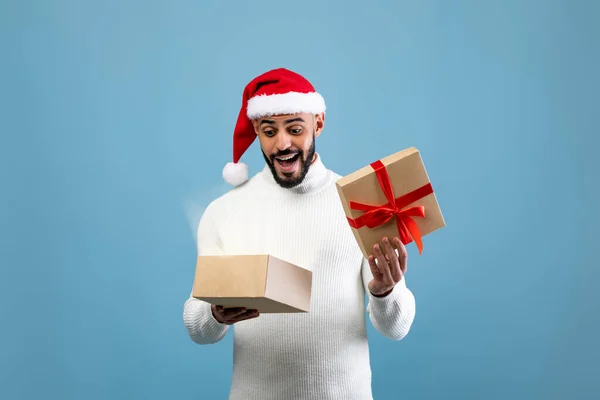 Milagro de Navidad. Hombre árabe excitado en Santa sombrero apertura envuelto caja de regalo y mirando dentro, fondo azul — Foto de Stock