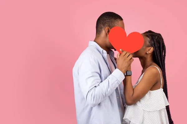 Jong romantisch zwart paar kussen verschuilen achter rood papier hart — Stockfoto