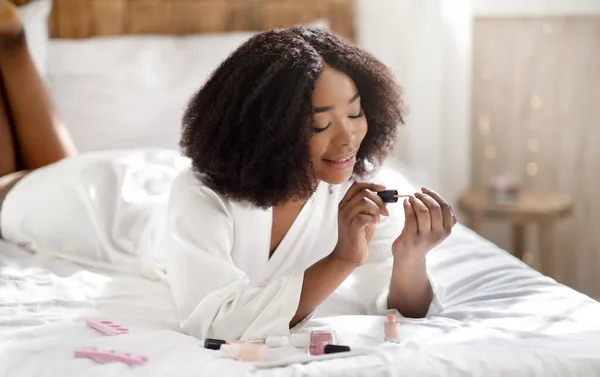 Mulher afro-americana feliz aplicando esmalte, fazendo manicure, deitado na cama, fazendo procedimento de spa doméstico — Fotografia de Stock