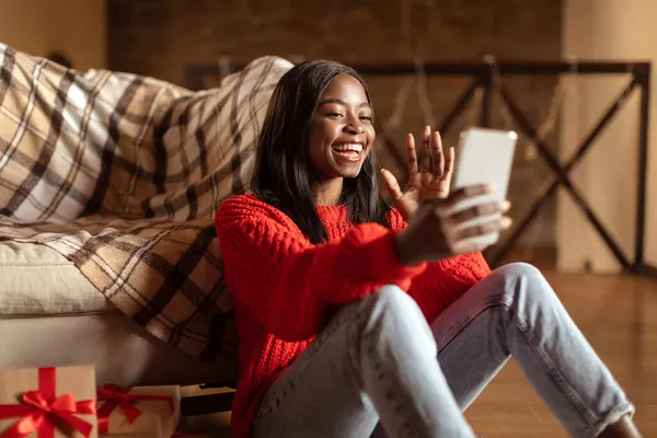 Fröhliche junge schwarze Dame im Winteroutfit winkt in die Smartphone-Kamera und plaudert mit Freund zu Hause — Stockfoto