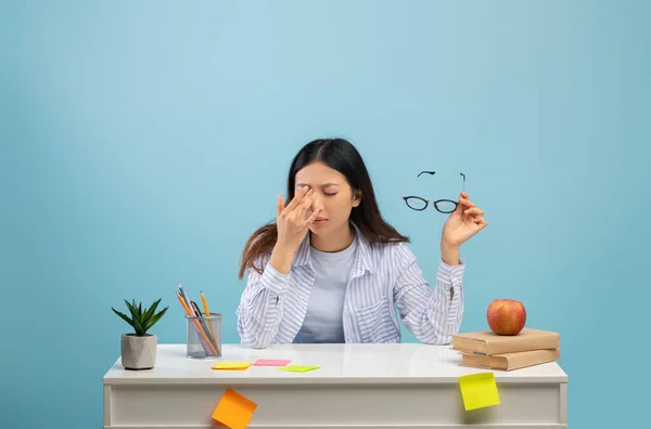 Zmęczona azjatycka pani masująca suche podrażnione oczy podczas nauki, przygotowująca się do egzaminów, siedząca przy biurku na niebieskim tle — Zdjęcie stockowe
