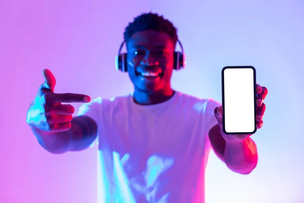Mira la nueva aplicación de música. Chico negro feliz en auriculares apuntando al teléfono inteligente con pantalla vacía en luz de neón, maqueta —  Fotos de Stock