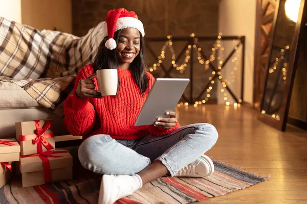 仮想Xmas 。サンタハットの黒人女性はコーヒーを飲み、タブレットを使用して家族とビデオ通話を行い、オンラインショッピング — ストック写真