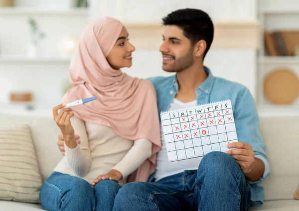 Planning pregnancy and expecting baby concept. Pretty arab couple holding positive pregnancy test and ovulation calendar