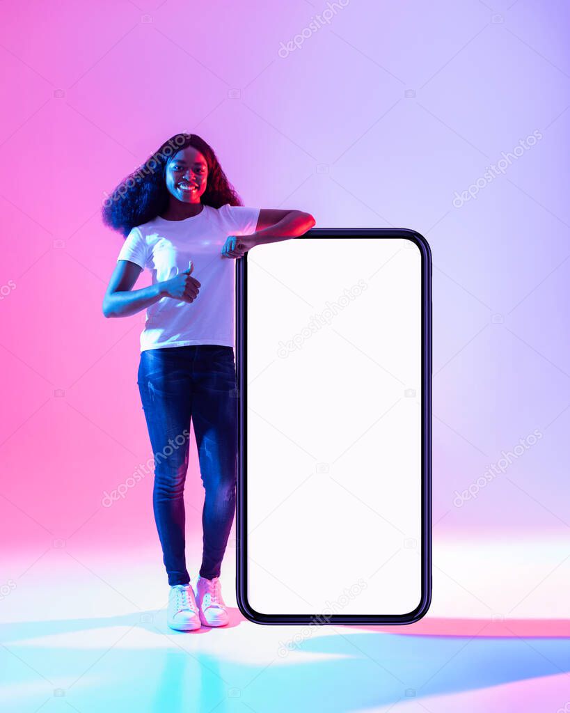 Happy black woman standing near big smartphone with blank white screen, gesturing thumb up in neon light, mockup