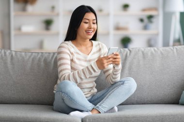 Evde koltukta dinlenirken akıllı telefon kullanan genç Asyalı kadın.
