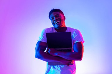 Teknoloji aşığı kavramı. Mutlu siyah adam dizüstü bilgisayarı boş ekranla sarılıyor neon ışığı altında, web sitesi tasarımı için model alıyor.