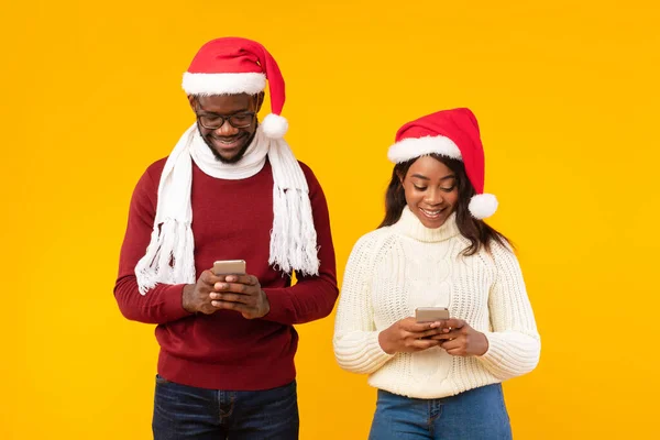 Black Couple Using Smartphones Wearing Santa Hats Over Yellow Background — Stock Photo, Image