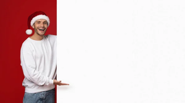 Weihnachtsliste. Fröhlicher Mann mit Weihnachtsmütze zeigt auf leere Werbetafel — Stockfoto