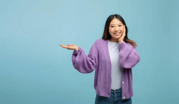 Wow, cooles Angebot. Fröhliche asiatische Dame zeigt leeren Raum auf Handfläche und rührende Wange vor blauem Hintergrund — Stockfoto