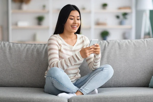 Ung asiatisk kvinna som använder smartphone medan du vilar på soffan hemma — Stockfoto