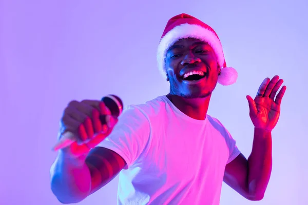 Weihnachtsmusik. Cooler schwarzer Kerl mit Weihnachtsmütze singt Weihnachtslied, benutzt Mikrofon, führt Karaoke im Neonlicht vor — Stockfoto