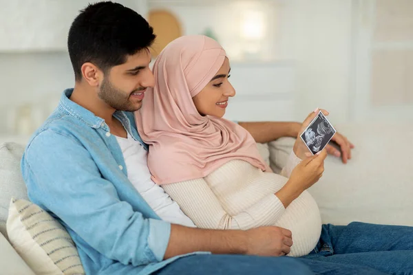 Čekám dítě. Nadšení těhotné muslimské rodiny při pohledu na ultrazvukové fotografie při odpočinku na pohovce, volný prostor — Stock fotografie