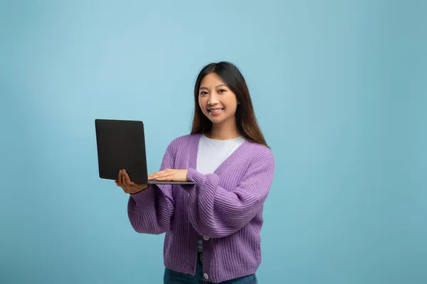 Porträt einer glücklichen jungen asiatischen Dame, die einen Laptop über blauem Studiohintergrund hält und in die Kamera lächelt — Stockfoto
