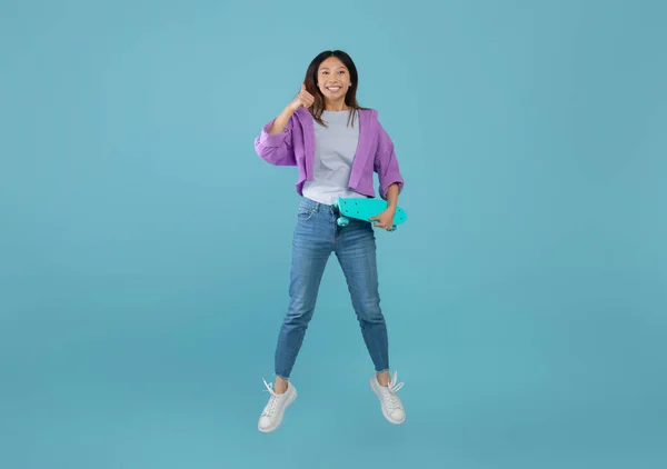 Mám rád teenagerskou kulturu. Radostné asijské dáma s skateboard jumping a ukazující palec nahoru přes modré pozadí — Stock fotografie