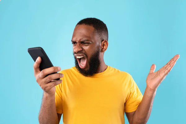 Θυμωμένος νεαρός μαύρος να φωνάζει στο smartphone πάνω από το μπλε φόντο του στούντιο. Διαχείριση άγχους και αρνητικά συναισθήματα — Φωτογραφία Αρχείου