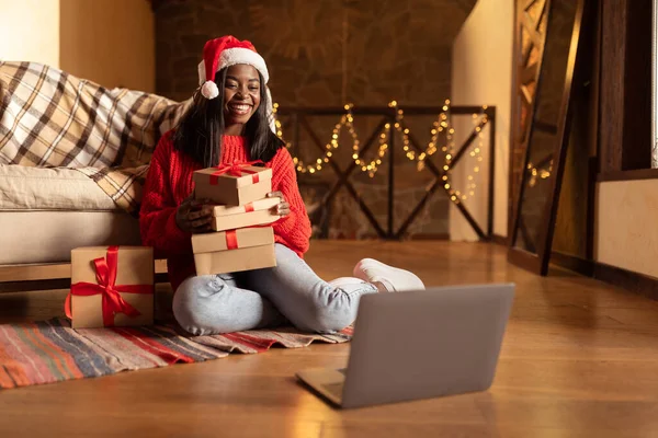 Giovane donna nera che tiene scatole regalo di Natale davanti al computer portatile, congratulandosi con la famiglia con le vacanze di Natale a casa — Foto Stock