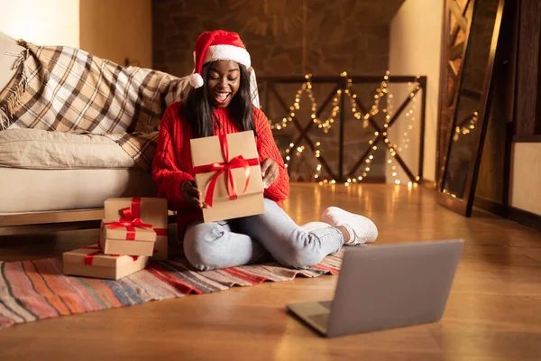 Excited black woman opening Christmas gifts in front of laptop, celebrating Xmas or New Year online at home, copy space