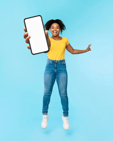 Gran nueva aplicación móvil. Emocionado mujer afroamericana saltando y mostrando teléfono inteligente con pantalla en blanco, maqueta — Foto de Stock