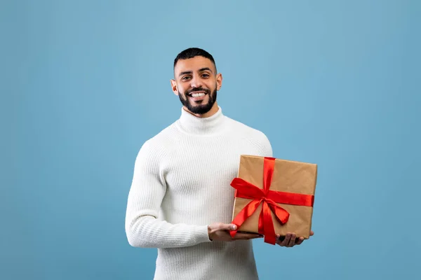 Senin için en iyi hediye. Mutlu genç Arap adam hediye kutusunu tutuyor, kameraya gülümsüyor, mavi arka planda duruyor. — Stok fotoğraf
