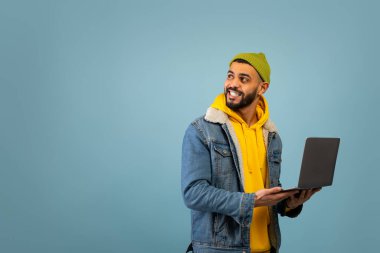 Uzaktan kumanda. Dizüstü bilgisayarlı mutlu genç Arap adam boş alana bakıyor, mavi arka planda duruyor.