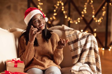 Noel Baba şapkalı mutlu siyahi bayan koltukta oturuyor, Noel arifesinde cep telefonuyla konuşuyor, evde boş yer var.