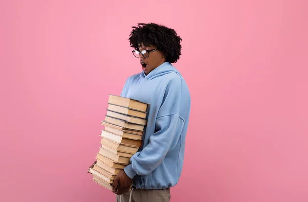 Overwhelmed black teenage student holding big stack of books, having lots of homework on pink background