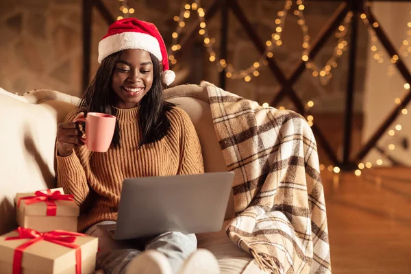 Porträtt av ung svart kvinna i Santa hatt dricka kaffe, handla julklappar online på bärbar dator hemma — Stockfoto