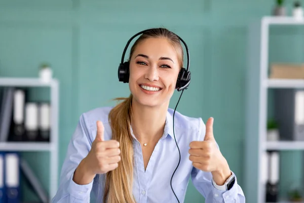 Távoktatás, munka. Boldog hölgy fülhallgató mutatja hüvelykujját fel gesztus, nézi és mosolyog a kamera az irodában — Stock Fotó