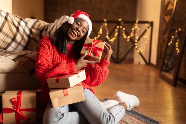 Noël et Nouvel An. Joyeux jeune femme noire dans le chapeau de Père Noël tenant cadeaux de Noël, assis sur le sol à la maison — Photo