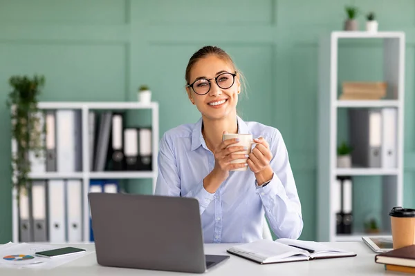 Goede dag op het werk. Gelukkig vrouwelijke kantoormedewerker drinken van koffie op de werkplek, werken op laptop computer, glimlachen op de camera — Stockfoto