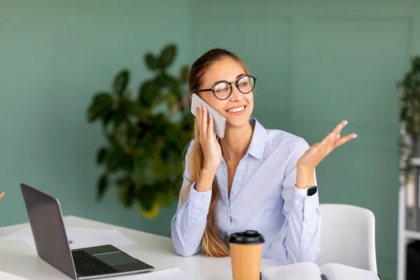 Comunicação corporativa. Empresária feliz falando no celular enquanto trabalhava no laptop no interior do escritório moderno — Fotografia de Stock