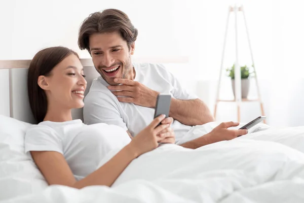 Mosolygó európai férfi és nő nézi a telefonokat, együtt nevetnek, ágyban fekszenek a hálószobában, szabad hely — Stock Fotó