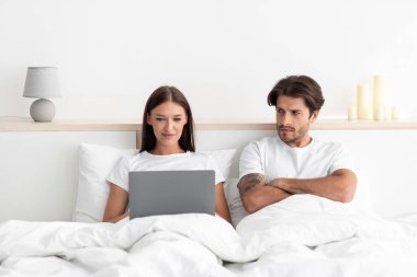 Huysuz, üzgün, gücenmiş, kollarını kavuşturmuş beyaz bir koca karısına bakar ve dizüstü bilgisayarında yazar.