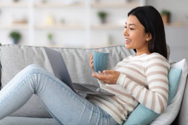 Asyalı kadın bilgisayarla video görüşmesi yapıyor ve evde kahve içiyor.