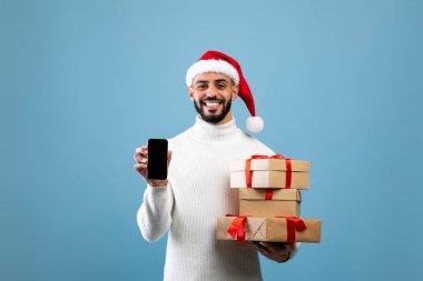 Noel reklamı için maket. Mutlu Arap adam, elinde hediyeler, mavi arka plan ve boş ekranlı akıllı telefon gösteriyor.